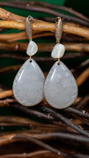 A pair of elegant teardrop-shaped earrings hanging on intertwined natural wooden branches. The earrings have a smooth, lustrous appearance with a subtly shimmering texture. Above each teardrop is a smaller, irregularly shaped bead, and both hang from gold-colored hooks with a rough, textured finish.