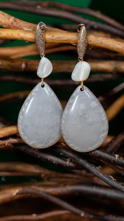 A pair of elegant teardrop-shaped earrings hanging on intertwined natural wooden branches. The earrings have a smooth, lustrous appearance with a subtly shimmering texture. Above each teardrop is a smaller, irregularly shaped bead, and both hang from gold-colored hooks with a rough, textured finish.