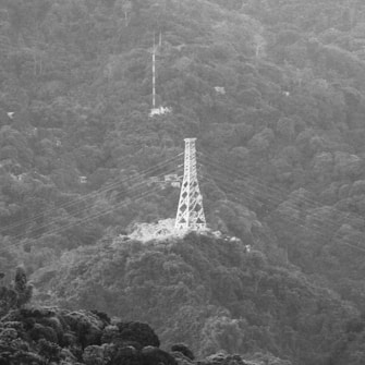 A transmission tower stands prominently on a heavily forested mountain slope, with power lines extending across the frame. The background is filled with dense foliage and uneven terrain. The overall grayscale tone gives a serene and timeless quality to the scene.