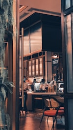 A cozy indoor restaurant setting with warm lighting and modern decor. The space features a counter where chefs in white uniforms and masks are working. There are jars of ingredients and bowls near the counter, and a sign with 'PASTA' is visible. A chair with a red cushion suggests it is a dining area.