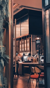 A cozy indoor restaurant setting with warm lighting and modern decor. The space features a counter where chefs in white uniforms and masks are working. There are jars of ingredients and bowls near the counter, and a sign with 'PASTA' is visible. A chair with a red cushion suggests it is a dining area.