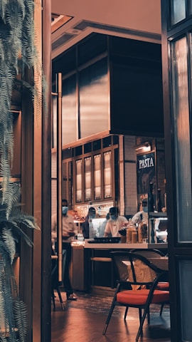 A cozy indoor restaurant setting with warm lighting and modern decor. The space features a counter where chefs in white uniforms and masks are working. There are jars of ingredients and bowls near the counter, and a sign with 'PASTA' is visible. A chair with a red cushion suggests it is a dining area.