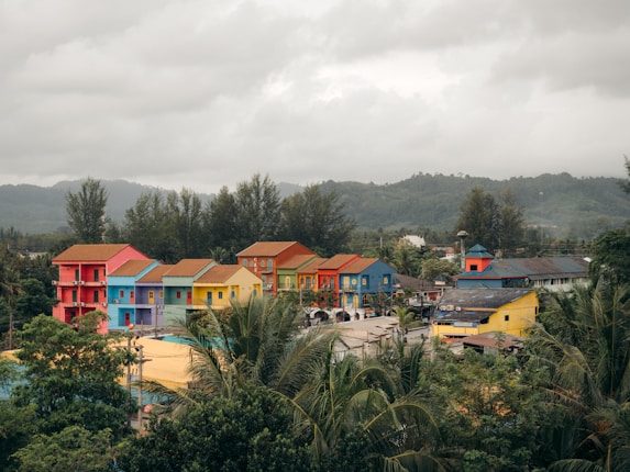 Colorful houses with vibrant hues of red, green, blue, yellow, and pink are arranged in a row. The setting is surrounded by lush greenery with mountains in the background under a cloudy sky.