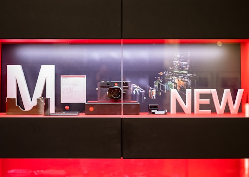 A display case contains a modern camera setup, showcased with large letters spelling 'M' and 'NEW'. The background illustrates a digital schematic of camera components. The interior of the display features an elegant, minimalistic arrangement with product cards and accessories.