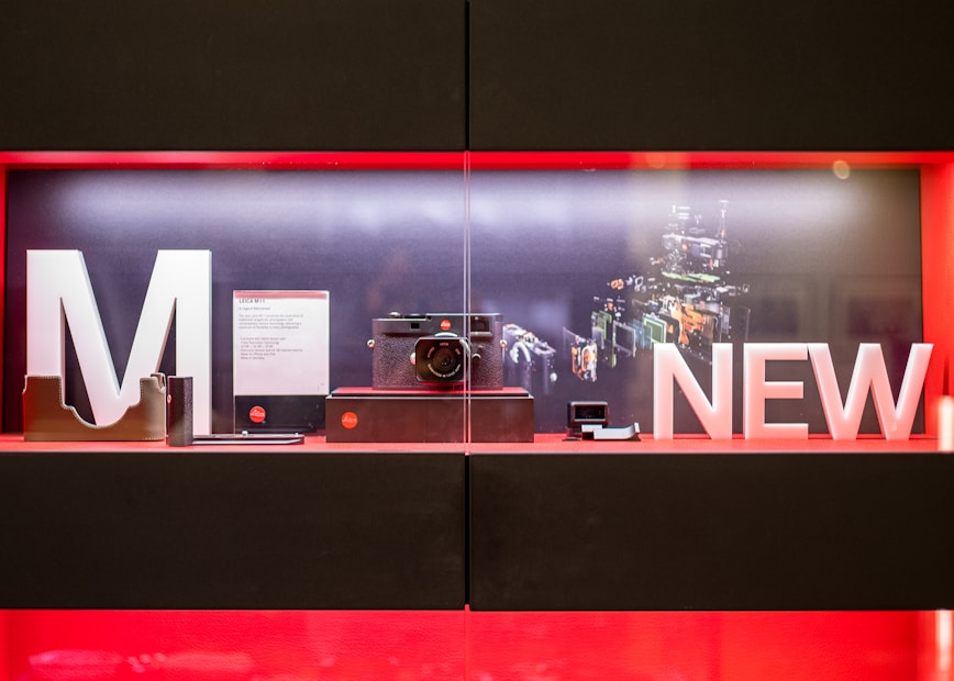 A display case contains a modern camera setup, showcased with large letters spelling 'M' and 'NEW'. The background illustrates a digital schematic of camera components. The interior of the display features an elegant, minimalistic arrangement with product cards and accessories.