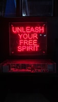 A red neon sign with the words 'Unleash Your Free Spirit' is prominently displayed above a dimly lit area, possibly a showcase or display. The bright neon contrasts with the dark background, creating a striking visual effect.
