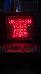 A red neon sign with the words 'Unleash Your Free Spirit' is prominently displayed above a dimly lit area, possibly a showcase or display. The bright neon contrasts with the dark background, creating a striking visual effect.