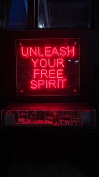 A red neon sign with the words 'Unleash Your Free Spirit' is prominently displayed above a dimly lit area, possibly a showcase or display. The bright neon contrasts with the dark background, creating a striking visual effect.