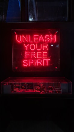 A red neon sign with the words 'Unleash Your Free Spirit' is prominently displayed above a dimly lit area, possibly a showcase or display. The bright neon contrasts with the dark background, creating a striking visual effect.