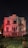 An old, dilapidated mansion stands in a field of overgrown grass, illuminated by moody red lights against a dark night sky. The building shows significant wear and decay, with peeling paint and broken windows, conveying a sense of abandonment.