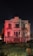 An old, dilapidated mansion stands in a field of overgrown grass, illuminated by moody red lights against a dark night sky. The building shows significant wear and decay, with peeling paint and broken windows, conveying a sense of abandonment.