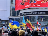A large group of people gather outside the Toronto Eaton Centre, holding various flags including Ukrainian, Lithuanian, and Canadian flags. The crowd is dressed warmly, suggesting cold weather. Above the crowd is a large digital billboard displaying promotional content with faces and text.
