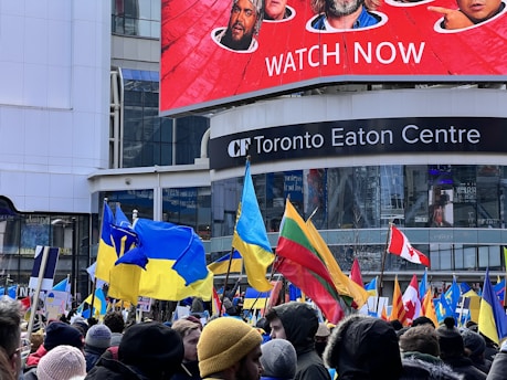 A large group of people gather outside the Toronto Eaton Centre, holding various flags including Ukrainian, Lithuanian, and Canadian flags. The crowd is dressed warmly, suggesting cold weather. Above the crowd is a large digital billboard displaying promotional content with faces and text.