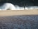 A person stands on a sandy beach, holding a fishing rod, facing a large wave crashing onto the shore. The foreground features a body of water with rippling effects.