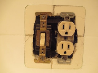 An exposed electrical outlet box featuring a light switch and a set of electrical sockets on a white wall. The surrounding wall appears to be unfinished, showing some paint and plaster inconsistencies. The metal components and screws are visible, with a slightly worn look.