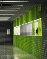 A hallway with modern architecture featuring green and dark gray tiled walls and floor. The lighting is bright and highlights the glossy surface of the tiles. A white framed picture hangs on the dark gray wall, and there is a window with a grid-like design on the green wall.