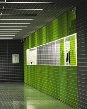 A hallway with modern architecture featuring green and dark gray tiled walls and floor. The lighting is bright and highlights the glossy surface of the tiles. A white framed picture hangs on the dark gray wall, and there is a window with a grid-like design on the green wall.