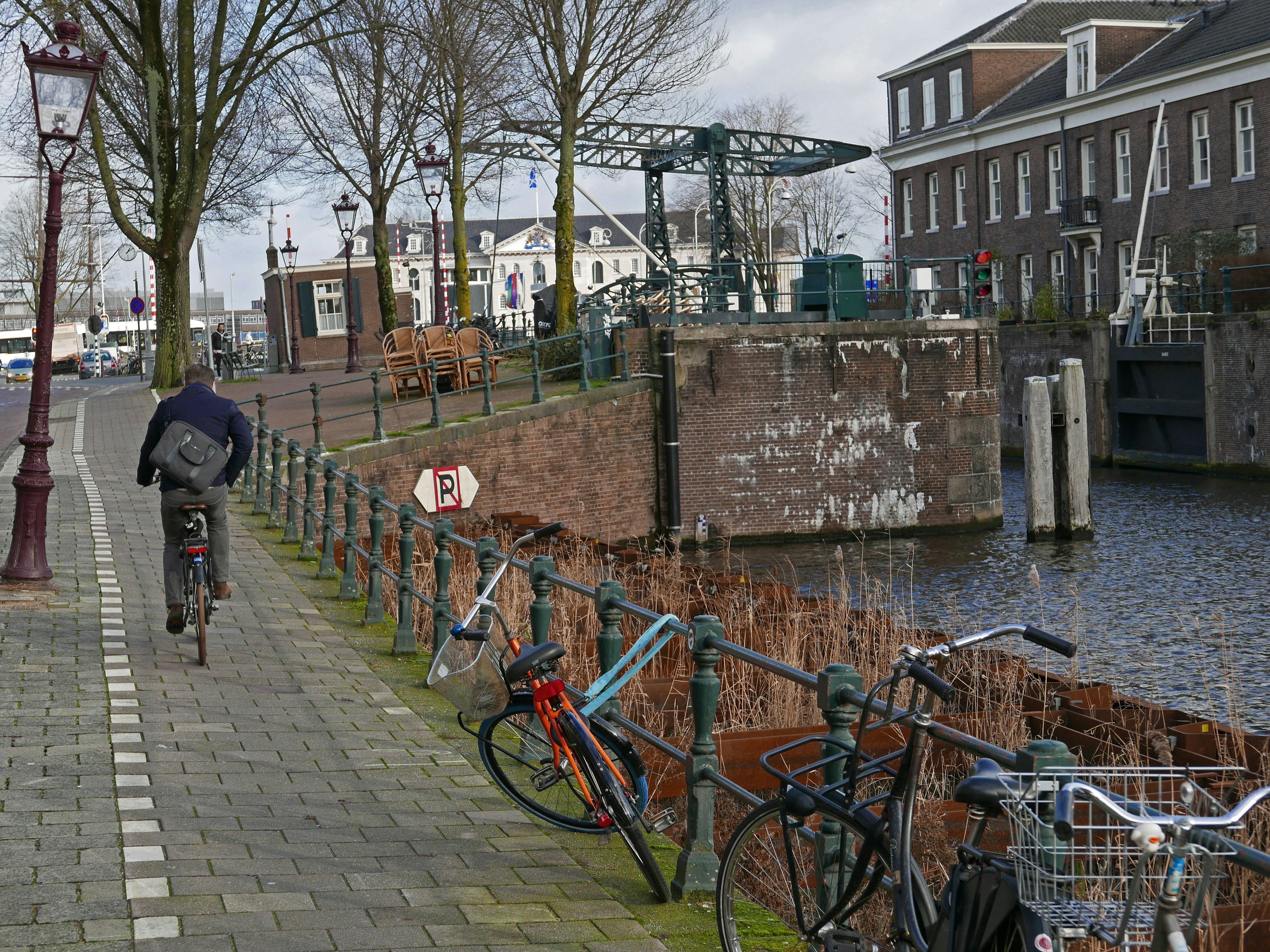 Street view with biker over the historical street Nieuwe Herengracht in Amsterdam city, along the canal water. To the right is an historic brick water lock, Picture of Amsterdam old canals - Dutch street photography in free pic photo by Fons Heijnsbroek, Netherlands. Foto van man / fietser op het fietspad bij oude sluis de Scharrebiersluis aan het einde van de Nieuwe Herengracht met metalen balans-brug over het kanaal. Op de achtergrond het Scheepvaartmuseum. Gratis download foto, Fons Heijnsbroek - fotografie van oude stad en historische grachten in Nederland, rechtenvrije afbeelding CCO