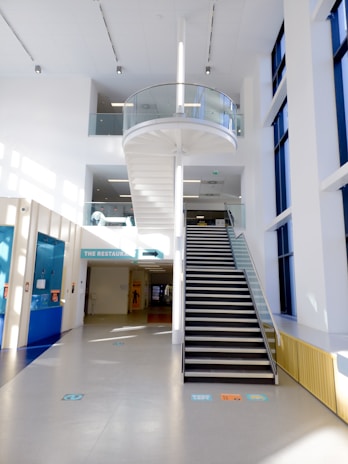 A modern interior space featuring a central staircase with glass railing leading to an upper level. The area is well-lit with natural light streaming through tall windows on the right. A sign overhead indicates the direction to a restaurant. The flooring is sleek with minimalistic design elements visible throughout.