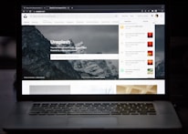A laptop displaying a website with an image of mountains in the background. The site is Unsplash, as indicated by the title. There are tabs and notifications on the right side and search functionality below the site’s name. The surroundings are dark, highlighting the screen as the primary light source.