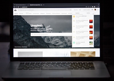 A laptop displaying a website with an image of mountains in the background. The site is Unsplash, as indicated by the title. There are tabs and notifications on the right side and search functionality below the site’s name. The surroundings are dark, highlighting the screen as the primary light source.