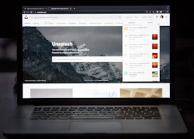 A laptop displaying a website with an image of mountains in the background. The site is Unsplash, as indicated by the title. There are tabs and notifications on the right side and search functionality below the site’s name. The surroundings are dark, highlighting the screen as the primary light source.