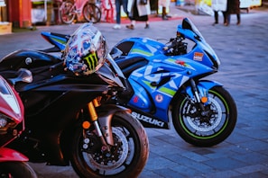 A close-up shot of three high-performance motorcycles parked on a paved surface. The primary focus is on two bikes: a sleek black Yamaha with a colorful racing helmet resting on its handlebars, and a vibrant blue Suzuki with racing decals and branding. In the background, there are people walking and bicycles, adding a hint of street life to the scene.