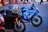 A close-up shot of three high-performance motorcycles parked on a paved surface. The primary focus is on two bikes: a sleek black Yamaha with a colorful racing helmet resting on its handlebars, and a vibrant blue Suzuki with racing decals and branding. In the background, there are people walking and bicycles, adding a hint of street life to the scene.