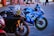 A close-up shot of three high-performance motorcycles parked on a paved surface. The primary focus is on two bikes: a sleek black Yamaha with a colorful racing helmet resting on its handlebars, and a vibrant blue Suzuki with racing decals and branding. In the background, there are people walking and bicycles, adding a hint of street life to the scene.