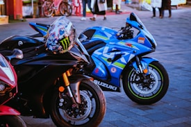 A close-up shot of three high-performance motorcycles parked on a paved surface. The primary focus is on two bikes: a sleek black Yamaha with a colorful racing helmet resting on its handlebars, and a vibrant blue Suzuki with racing decals and branding. In the background, there are people walking and bicycles, adding a hint of street life to the scene.
