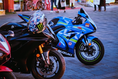 A close-up shot of three high-performance motorcycles parked on a paved surface. The primary focus is on two bikes: a sleek black Yamaha with a colorful racing helmet resting on its handlebars, and a vibrant blue Suzuki with racing decals and branding. In the background, there are people walking and bicycles, adding a hint of street life to the scene.