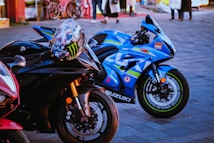 A close-up shot of three high-performance motorcycles parked on a paved surface. The primary focus is on two bikes: a sleek black Yamaha with a colorful racing helmet resting on its handlebars, and a vibrant blue Suzuki with racing decals and branding. In the background, there are people walking and bicycles, adding a hint of street life to the scene.