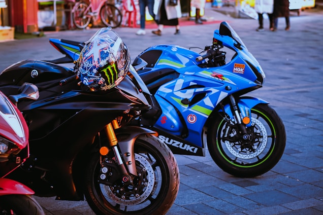 A close-up shot of three high-performance motorcycles parked on a paved surface. The primary focus is on two bikes: a sleek black Yamaha with a colorful racing helmet resting on its handlebars, and a vibrant blue Suzuki with racing decals and branding. In the background, there are people walking and bicycles, adding a hint of street life to the scene.