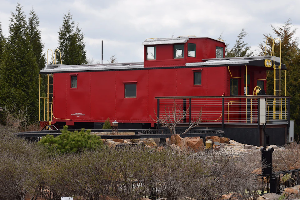 The image shows a red train car, possibly an old-fashioned steam locomotive or a modern caboose, parked on a track surrounded by trees and shrubs. The train car is painted in a striking shade of red with white trim, giving it a vintage appearance. It features two levels, one at the front and another at the back, suggesting that it could be used for passenger transportation or even as a mobile home.