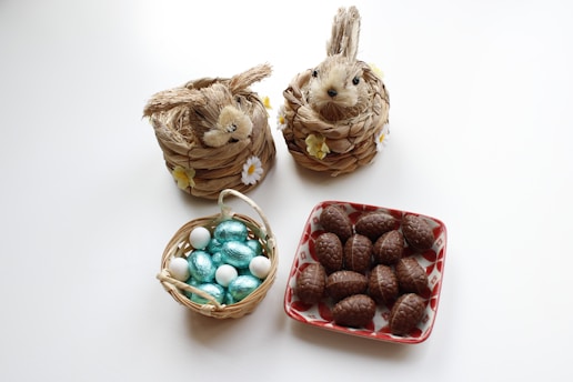 Two straw rabbit figures decorated with yellow and white flowers sit next to a small basket filled with turquoise and white foil-wrapped eggs. Nearby, a red and white patterned plate holds several chocolate egg-shaped treats.