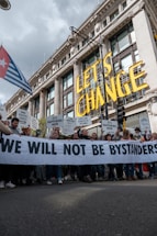 A large group of people are participating in a protest in front of a grand historic building. They are holding a banner that reads 'WE WILL NOT BE BYSTANDERS' and various signs with messages related to climate change and fossil fuels. The words 'LET'S CHANGE' are prominently displayed above the crowd.