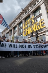 A large group of people are participating in a protest in front of a grand historic building. They are holding a banner that reads 'WE WILL NOT BE BYSTANDERS' and various signs with messages related to climate change and fossil fuels. The words 'LET'S CHANGE' are prominently displayed above the crowd.