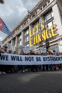 A large group of people are participating in a protest in front of a grand historic building. They are holding a banner that reads 'WE WILL NOT BE BYSTANDERS' and various signs with messages related to climate change and fossil fuels. The words 'LET'S CHANGE' are prominently displayed above the crowd.