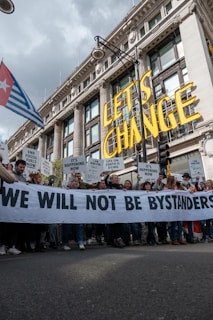 A large group of people are participating in a protest in front of a grand historic building. They are holding a banner that reads 'WE WILL NOT BE BYSTANDERS' and various signs with messages related to climate change and fossil fuels. The words 'LET'S CHANGE' are prominently displayed above the crowd.