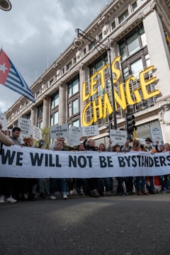 A large group of people are participating in a protest in front of a grand historic building. They are holding a banner that reads 'WE WILL NOT BE BYSTANDERS' and various signs with messages related to climate change and fossil fuels. The words 'LET'S CHANGE' are prominently displayed above the crowd.