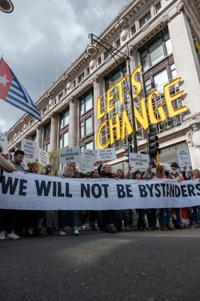 A large group of people are participating in a protest in front of a grand historic building. They are holding a banner that reads 'WE WILL NOT BE BYSTANDERS' and various signs with messages related to climate change and fossil fuels. The words 'LET'S CHANGE' are prominently displayed above the crowd.