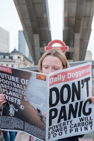 A person is holding a newspaper titled 'The Daily Doggerel' with headlines such as 'DON'T PANIC' and 'Poverty Will Lower Carbon Footprint.' The backdrop features what appears to be a London Underground sign and urban architecture.