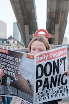 A person is holding a newspaper titled 'The Daily Doggerel' with headlines such as 'DON'T PANIC' and 'Poverty Will Lower Carbon Footprint.' The backdrop features what appears to be a London Underground sign and urban architecture.