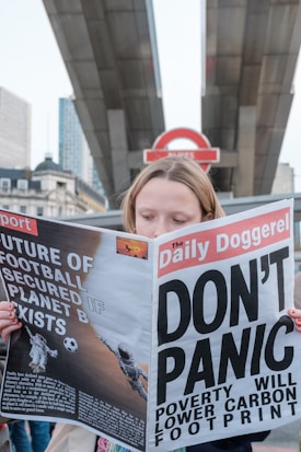 A person is holding a newspaper titled 'The Daily Doggerel' with headlines such as 'DON'T PANIC' and 'Poverty Will Lower Carbon Footprint.' The backdrop features what appears to be a London Underground sign and urban architecture.