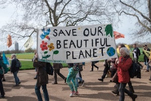A group of people are marching outdoors while holding a large banner with the message 'Save Our Beautiful Planet'. The banner is decorated with colorful drawings of flowers, leaves, and a small globe. Various flags are being carried, and there are trees without leaves in the background.