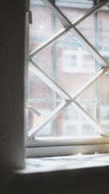 A window with a diamond-shaped grid pattern partially open, allowing a view of a blurry building facade in the background. The frame appears to be painted in a light color, possibly white, and the glass exhibits some dirt or condensation.