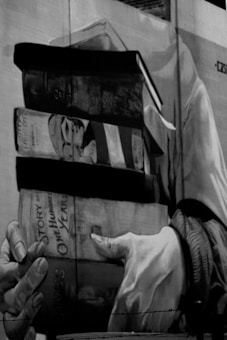 A monochrome mural features a pair of hands holding several large books. The titles include 'Gone with the Wind' and 'One Hundred Years of Solitude,' among others. The artwork appears to be painted on a large wall surface, giving prominence to the books and the detailed depiction of the hands.