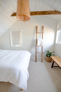 A minimalist bedroom features a white bed with a wooden frame, a rustic wooden ladder with a towel draped over it, a woven basket planter with a green plant, and a simple wooden bench. The ceiling has exposed wooden beams and a large wicker light fixture. The walls and ceiling are painted white, enhancing the bright and airy feel.