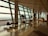 A spacious airport terminal with large glass windows letting in natural light, creating reflections on the polished floor. There are empty lounge chairs along the windows and a few potted plants adding greenery. A sign with the number 36 is visible next to a small desk area.