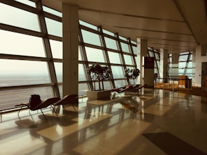 A spacious airport terminal with large glass windows letting in natural light, creating reflections on the polished floor. There are empty lounge chairs along the windows and a few potted plants adding greenery. A sign with the number 36 is visible next to a small desk area.